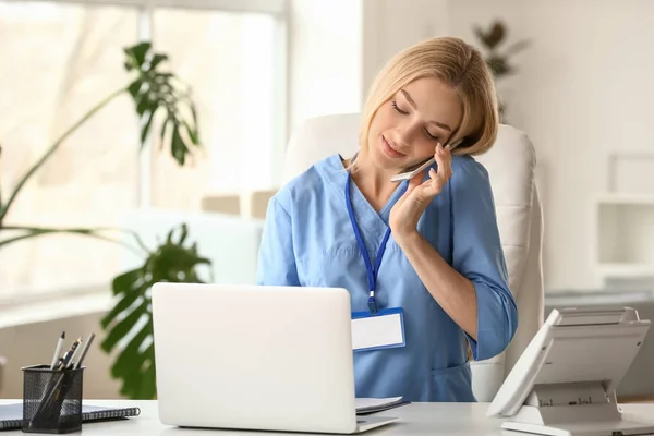 Female medical assistant talking by phone while working on laptop in clinic