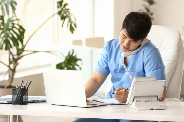Male medical assistant talking by phone while working on laptop in clinic — Stock Photo, Image