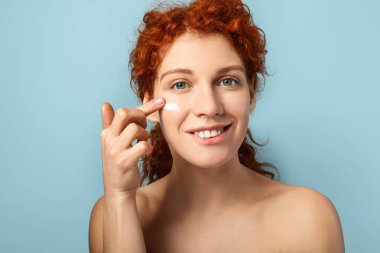 Beautiful redhead woman applying cream against color background