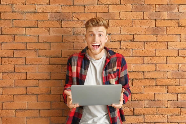 Surprised young man with laptop near brick wall — Stock Photo, Image