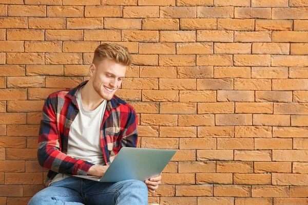 Handsome young man with laptop sitting near brick wall — Stock Photo, Image
