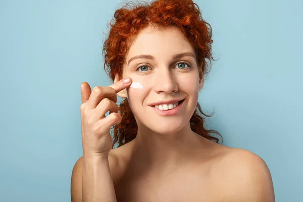 Hermosa mujer pelirroja aplicando crema contra el fondo de color — Foto de Stock