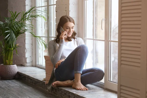 Gyönyörű fiatal nő beszél mobiltelefonos közelében ablak otthon — Stock Fotó