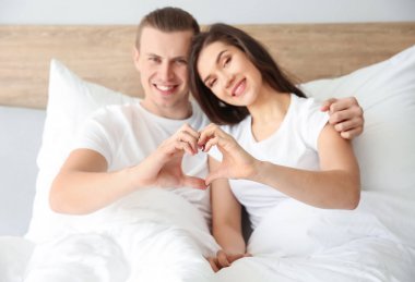 Yatakta otururken kalp şeklinde el ele tutuşan mutlu genç çift