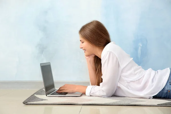 Beautiful young woman with laptop lying on floor against color wall — Stock Photo, Image