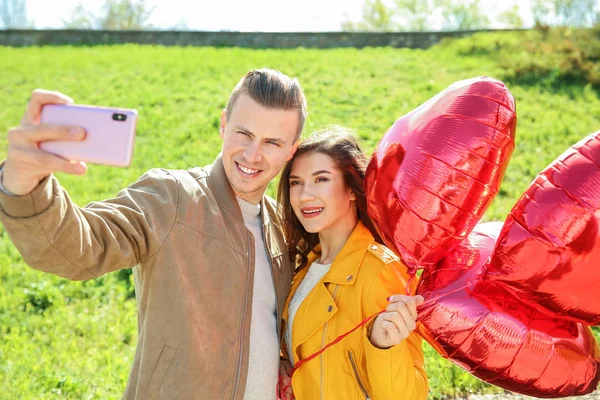 Feliz pareja joven con globos de aire en forma de corazón tomando selfie al aire libre — Foto de Stock