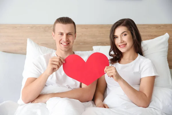Feliz jovem casal com papel coração sentado na cama — Fotografia de Stock