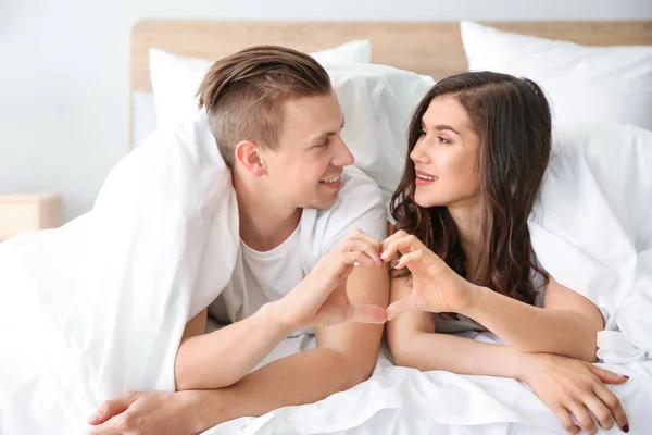 Feliz jovem casal de mãos dadas em forma de coração enquanto deitado na cama — Fotografia de Stock