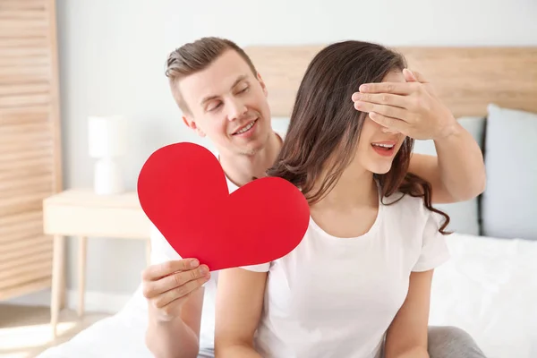 Joven con corazón de papel cubriendo los ojos de su esposa en casa — Foto de Stock