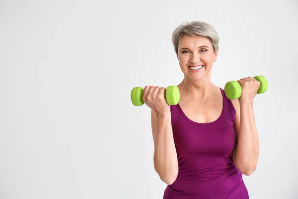 Sporty mature woman with dumbbells on light background — Stock Photo, Image