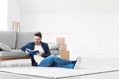 Genç adam evde yumuşak halıda otururken kitap okuyor.