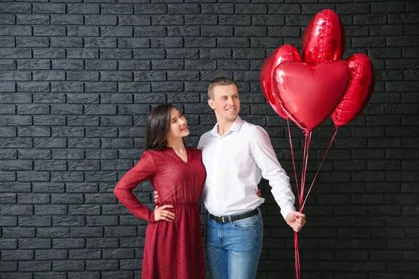 Feliz pareja joven con globos de aire cerca de la pared de ladrillo oscuro —  Fotos de Stock