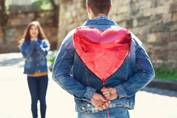 Jeune homme cachant ballon d'air en forme de coeur pour petite amie derrière son dos à l'extérieur — Photo