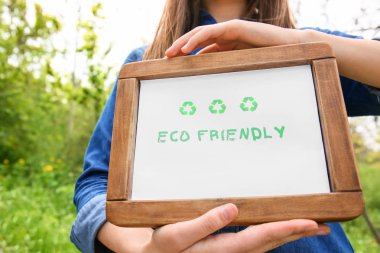 Woman holding board with text ECO FRIENDLY outdoors, closeup