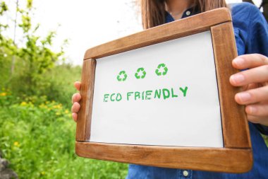 Woman holding board with text ECO FRIENDLY outdoors, closeup