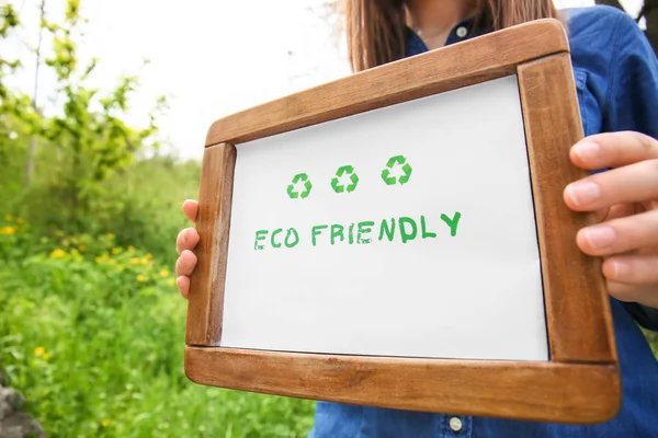 Woman holding board with text ECO FRIENDLY outdoors, closeup