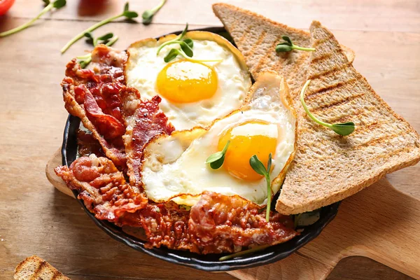 Frying pan with tasty eggs, bacon and toasts on table — Stock Photo, Image