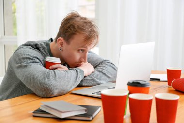 Yorgun adam dizüstü bilgisayar üzerinde çalışıyor ve evde kahve çok içme