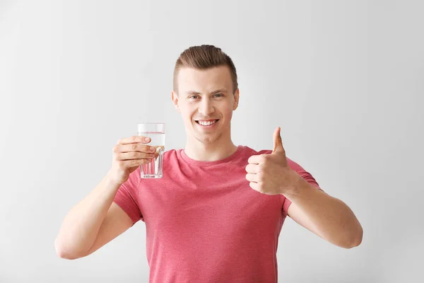 Knappe jonge man met glas water weergegeven duim-up op lichte achtergrond — Stockfoto