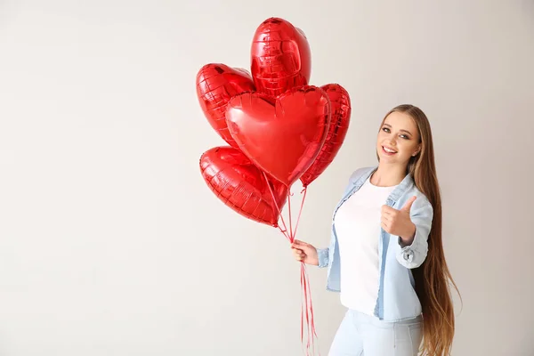 Beautiful young woman with heart-shaped air balloons showing thumb-up on light background — Stock Photo, Image