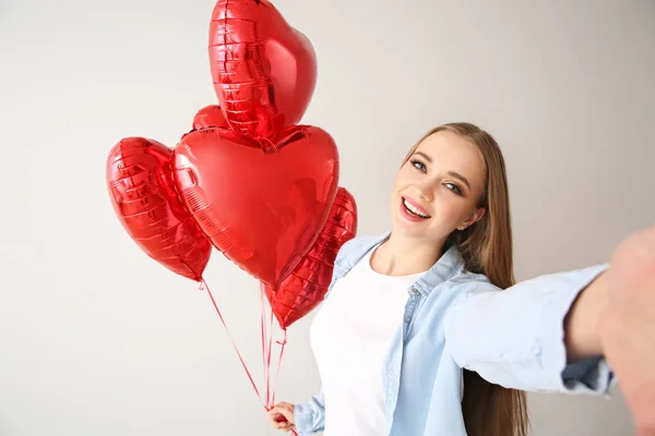 Mulher bonita com balões de ar em forma de coração tomando selfie no fundo claro — Fotografia de Stock