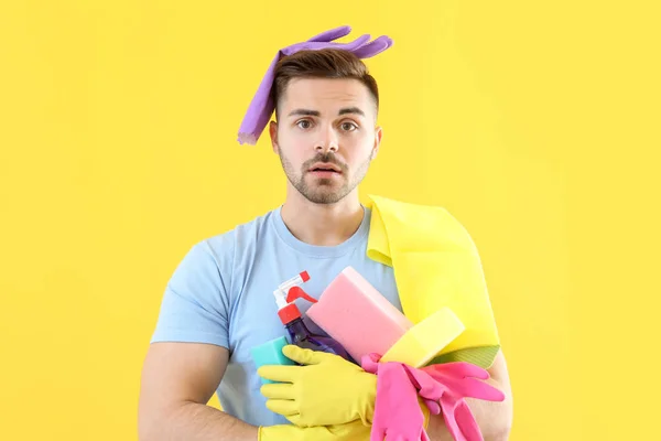 Stressed young man with cleaning supplies on color background