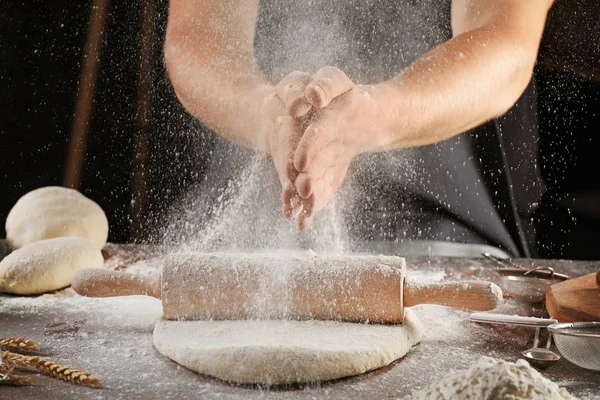 Hombre aplaudiendo las manos y rociando la harina sobre la masa en la mesa — Foto de Stock