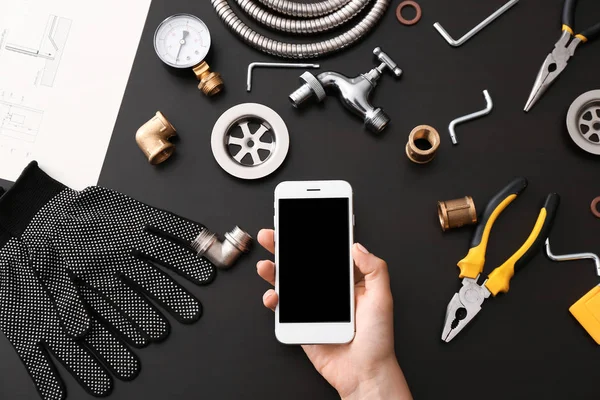 Plumber tools and female hand with mobile phone on dark background, top view