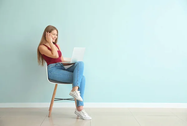 Beautiful young woman with laptop sitting on chair against color wall — Stock Photo, Image