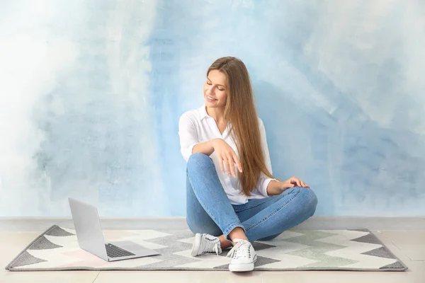 Beautiful young woman with laptop sitting on floor against color wall — Stock Photo, Image