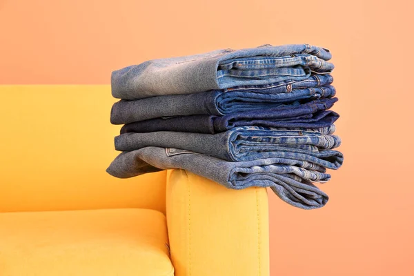 Stack of jeans pants on armchair against color background — Stock Photo, Image