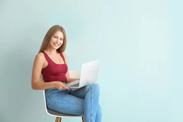 Beautiful young woman with laptop sitting on chair against color background — Stock Photo, Image