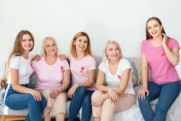 Hermosas mujeres de diferentes edades con cintas rosadas sentadas en el sofá en la habitación. Concepto de cáncer de mama — Foto de Stock