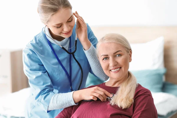 Medical assistant examining mature patient with stethoscope at home