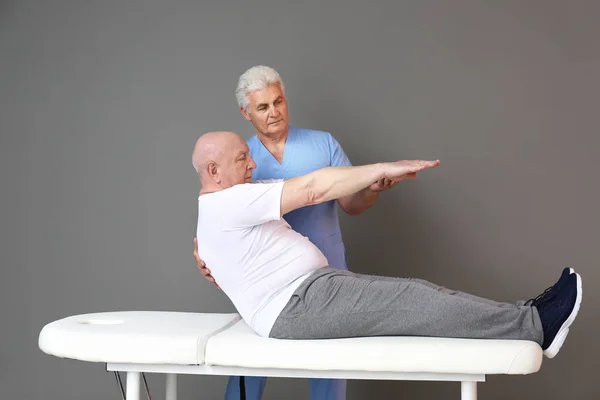 Зрелый физиотерапевт, работающий со старшим мужчиной на сером фоне — стоковое фото