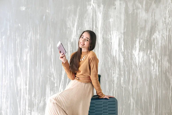 Šťastná mladá žena s kufříkem, pasem a letenkou poblíž šedé zdi — Stock fotografie
