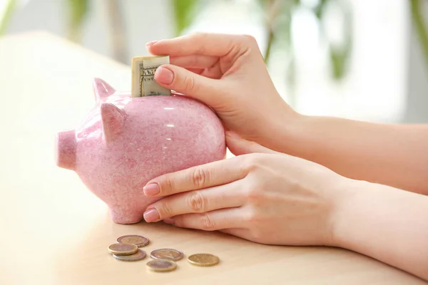 Young woman putting coins in piggy bank on table, closeup — Stock Photo, Image