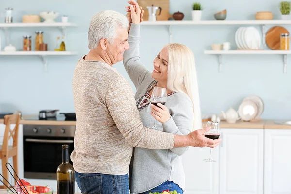 Retrato de pareja madura feliz bailando y bebiendo vino en la cocina —  Fotos de Stock