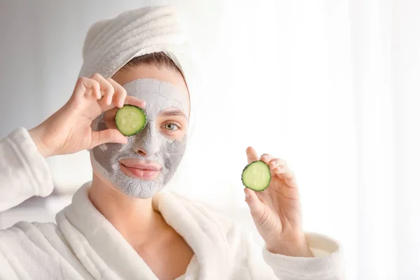 Jonge vrouw met klei masker en komkommer plakjes thuis — Stockfoto