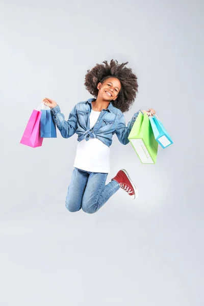 Jumping African-American girl in jeans clothes and with shopping bags on grey background — Stock Photo, Image