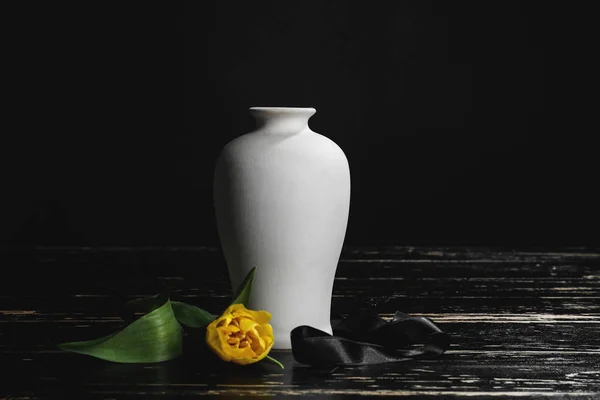 Urne mit Schleife und Blume auf Tisch vor dunklem Hintergrund — Stockfoto