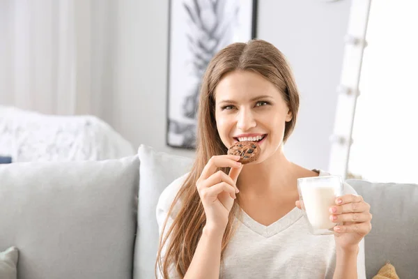 Beautiful young woman eating tasty cookie with milk at home — Stock Photo, Image