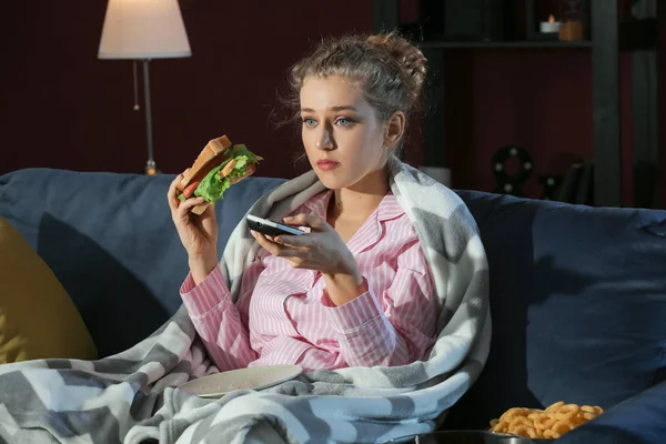 Hermosa mujer joven comiendo alimentos poco saludables mientras ve la televisión por la noche —  Fotos de Stock