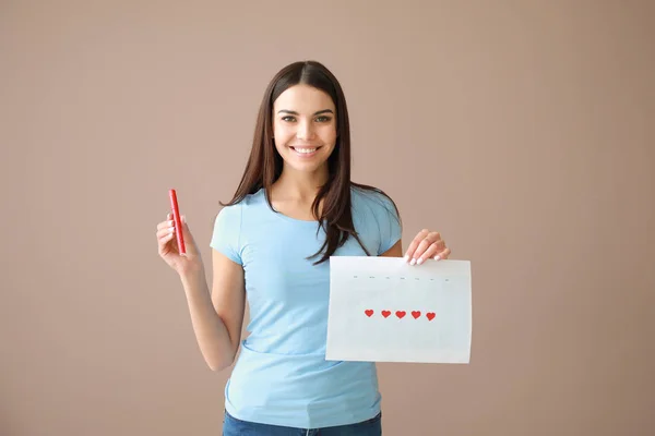 Young woman holding calendar with marked days of menstruation on color background — Stock Photo, Image