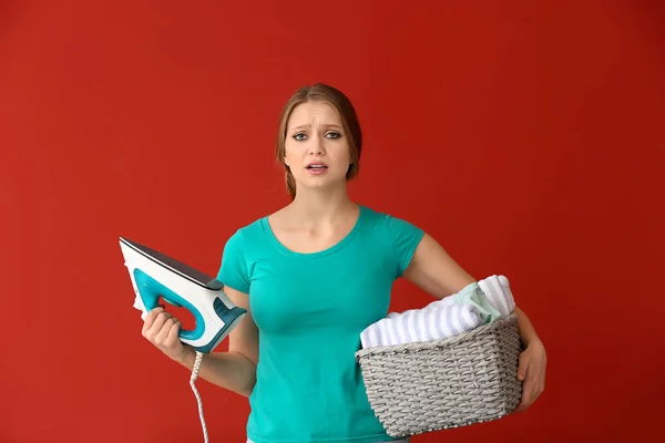 Sad young woman with iron and clean clothes on color background — Stock Photo, Image