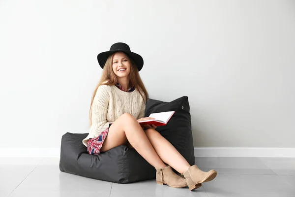 Hermosa joven con libro sentado cerca de la pared de luz —  Fotos de Stock