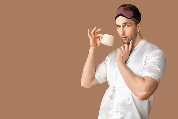 Funny young man with sleep mask and cup of coffee on color background — Stock Photo, Image