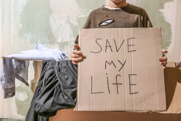 Niño sin hogar sosteniendo pedazo de cartón con texto Save My Life en interiores — Foto de Stock