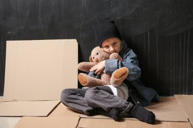 Karanlık duvarın yanında yerde oyuncak ayı oturan evsiz küçük kız