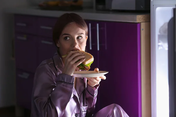 Mujer joven cautelosa comiendo alimentos poco saludables cerca del refrigerador por la noche —  Fotos de Stock